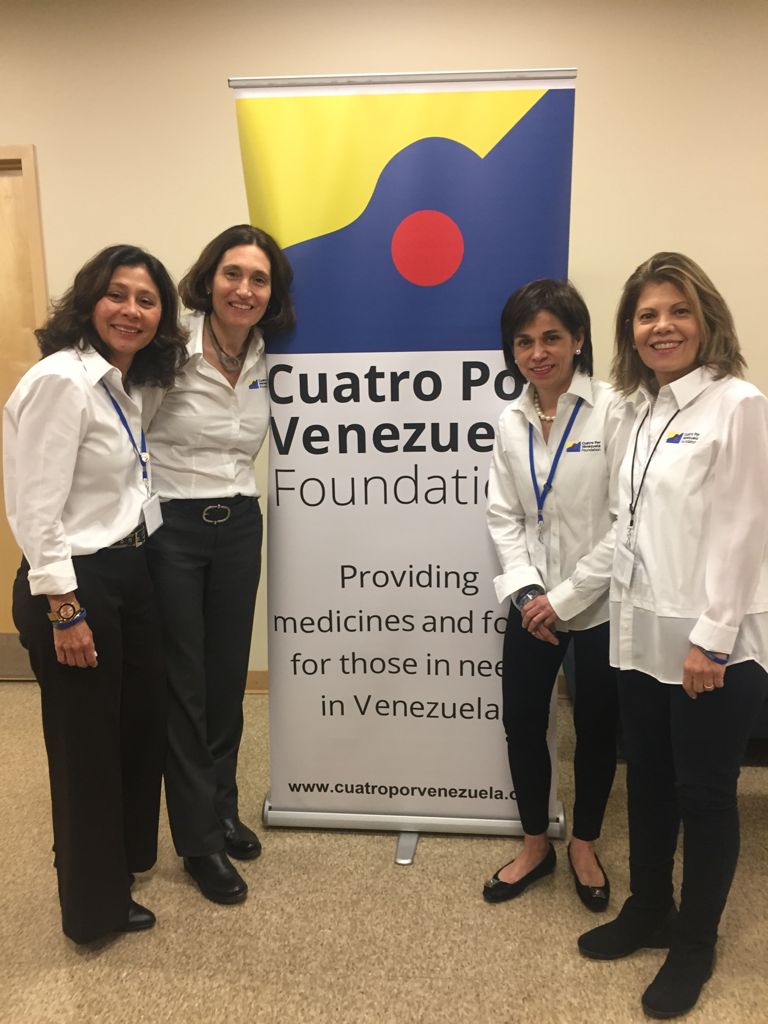 Photo of Four Women in White Shirts Standing by a Cuatro Por Venezuela Foundation Banner